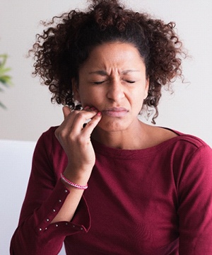 A middle-aged woman wearing a burgundy shirt and holding the side of her mouth because of intense pain and discomfort caused by gum disease