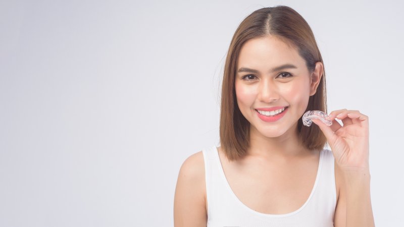 Young woman smiling with teeth and holding clear aligner up to her chin with her left hand